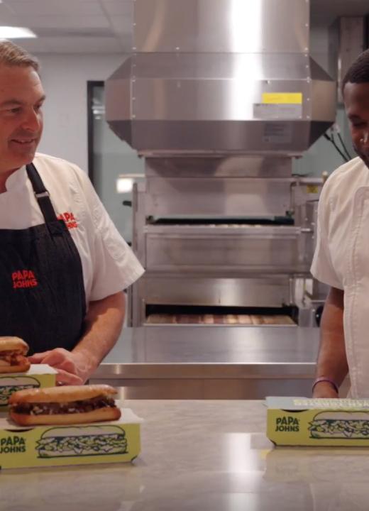 Two men standing at a table with sandwiches.