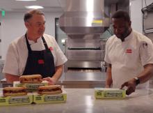 Two men standing at a table with sandwiches.