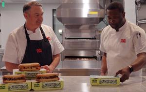 Two men standing at a table with sandwiches.