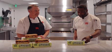 Two men standing at a table with sandwiches.