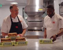 Two men standing at a table with sandwiches.